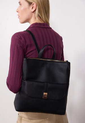 Frau mit blondem Haar, die ein lila Oberteil und beige Hosen trägt, und einen großen schwarzen Leder-Rucksack mit Fronttasche und goldener Schnalle trägt.