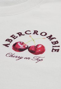 Λευκό ύφασμα με γραφικό σχέδιο που απεικονίζει δύο κόκκινα κεράσια και το κείμενο «Abercrombie Cherry on Top» σε σκούρα βυσσινί γράμματα.