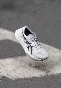 White athletic shoe with black accents, mesh upper, perforated texture, and lightweight sole, suspended above a pavement marked with white stripes.