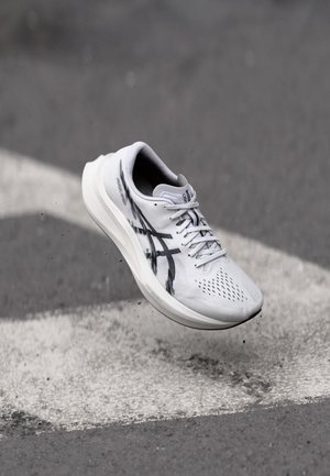 White athletic shoe with black accents, mesh upper, perforated texture, and lightweight sole, suspended above a pavement marked with white stripes.