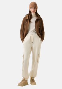 Brun ribbet zip-jakke, creme turtleneck, creme cargo-bukser med sidelommer og beige sneakers med teksturerede detaljer. Beige strikket beanie.