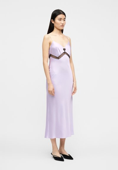 Γυναίκα που φοράει φόρεμα από σατέν λεβάντας με μαύρη δαντέλα στο τελείωμα και μαύρα pointed mules με χαμηλό τακούνι, στέκεται μπροστά σε ένα λευκό φόντο.