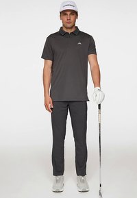 Grå pikétröja med krage och logotyp, kombinerad med matchande grå byxor. Bär vit golfhandske och sneakers. Står med en golfklubba.