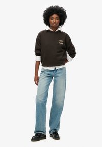 Felpa marrone con ricamo "UNION #6 Superdry", indossata sopra una camicia bianca con colletto. Jeani larghi blu chiaro, scarpe nere. Capelli afro.