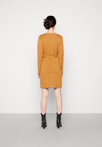Abito sweater color senape con maniche lunghe, vita aderente e dettaglio con fiocco sul retro, abbinato a stivaletti neri alla caviglia. Tessuto liscio, design minimale.
