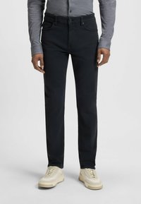 Pantalones de mezclilla negros con un ajuste entallado, cinco bolsillos y cierre de botón. Llevados con zapatillas crema y una camisa de manga larga gris.