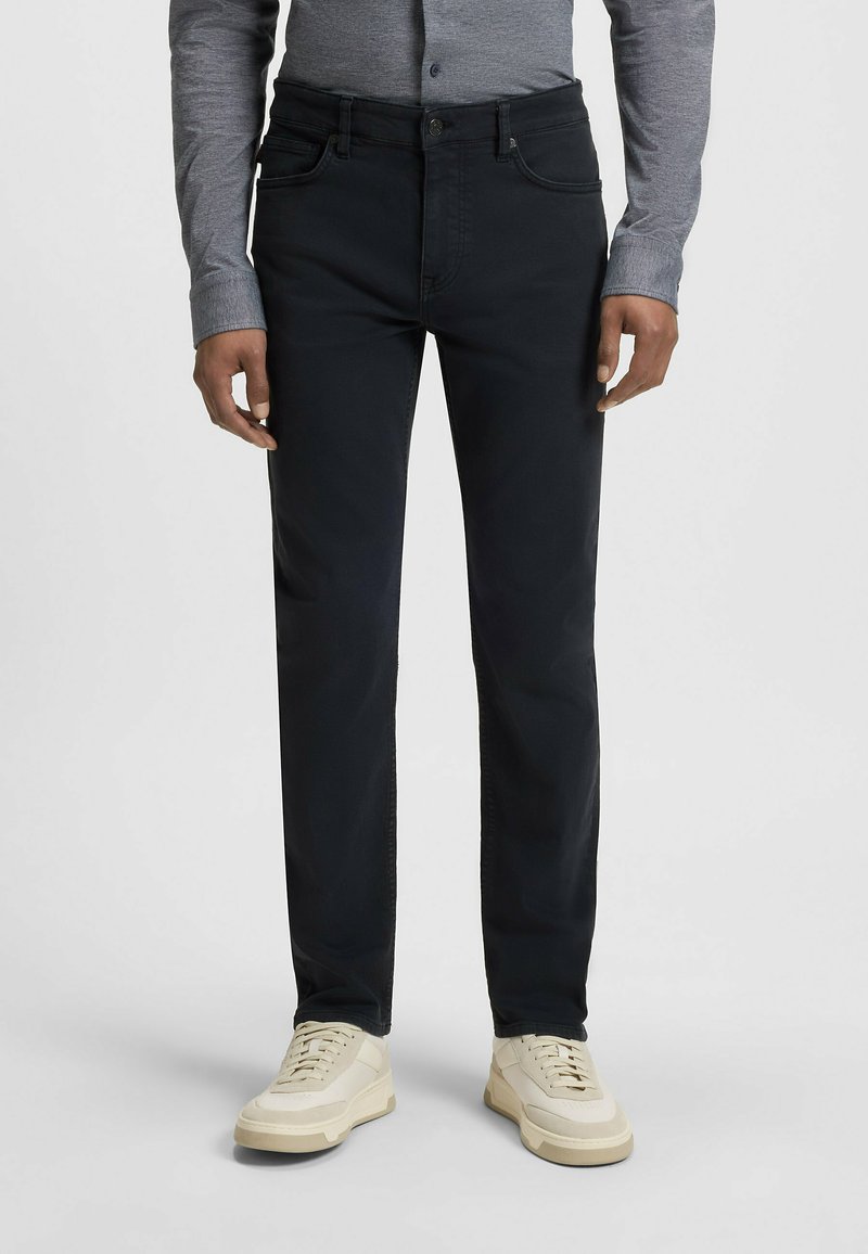 Pantalones de mezclilla negros con un ajuste entallado, cinco bolsillos y cierre de botón. Llevados con zapatillas crema y una camisa de manga larga gris.