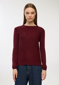 Bordowy sweter dzianinowy z okrągłym dekoltem i prążkowanymi mankietami, o miękkiej fakturze i klasycznym prostym kroju. Noszony z niebieskimi dżinsami.