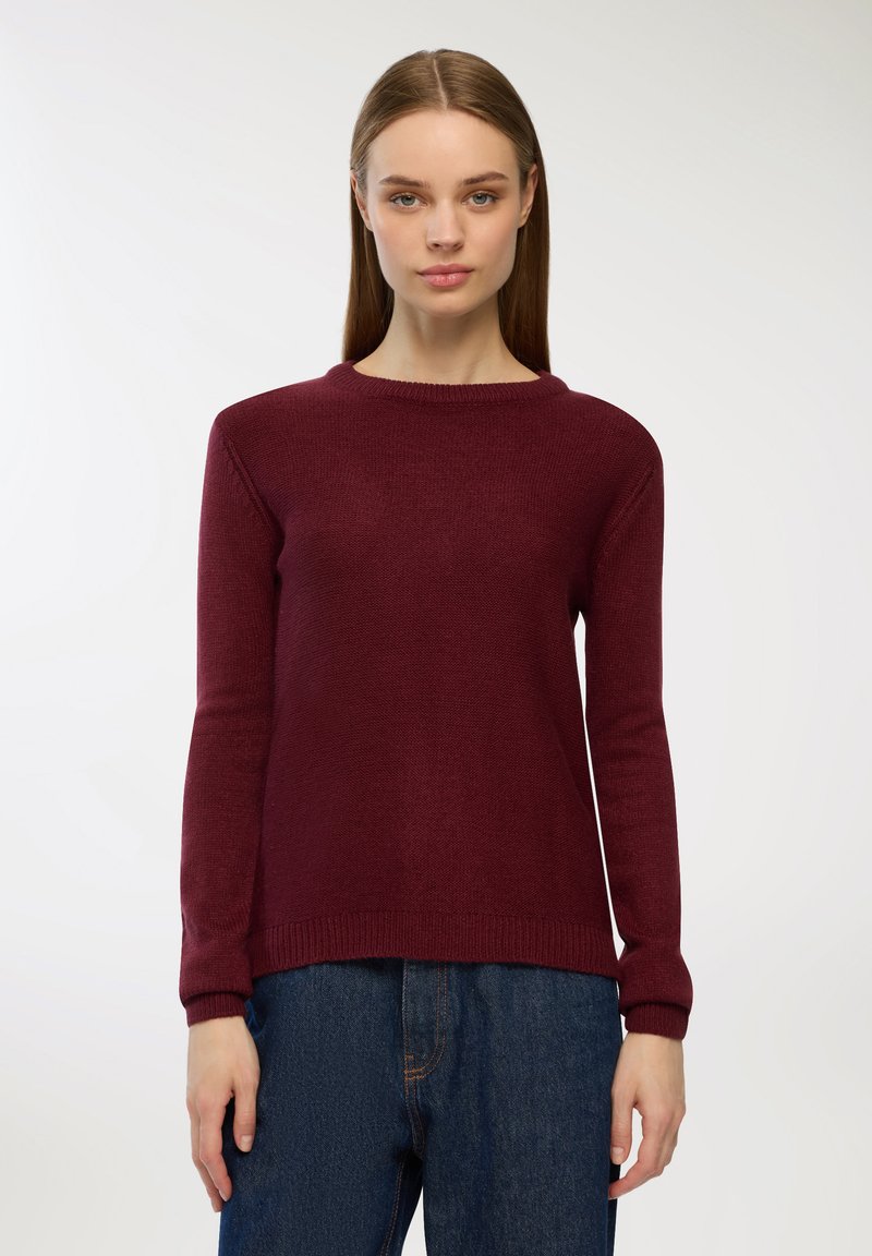 Bordowy sweter dzianinowy z okrągłym dekoltem i prążkowanymi mankietami, o miękkiej fakturze i klasycznym prostym kroju. Noszony z niebieskimi dżinsami.