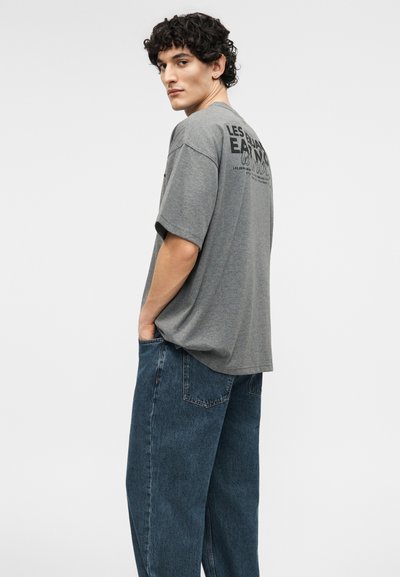 Joven con cabello rizado y oscuro, vistiendo una camiseta gris holgada con texto negro en la parte trasera y jeans anchos de mezclilla azul, de pie con las manos en los bolsillos.