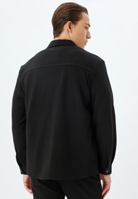 Maglietta nera a maniche lunghe con texture liscia, colletto e polsini con bottone. Presenta una cucitura orizzontale nella parte superiore della schiena per un dettaglio di design.