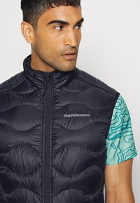 Jūras spēku pufīgā veste ar quilt dizainu, priekšējo rāvējslēdzēju un Peak Performance logo. Valkāta virs rakstainas īsām piedurknēm blūzes.