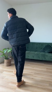 Črna puhovka s horizontalnim šivom, v kombinaciji s temnimi kavbojkami in svetlo rjavimi čevlji. Notranja postavitev z zeleno zofjo in rastlino.
