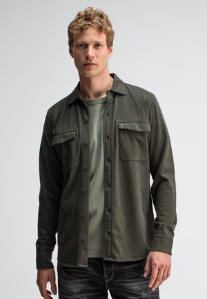 Olive green long-sleeve shirt with two front pockets, button-down front, and soft texture. Worn over a matching green T-shirt, paired with dark jeans.