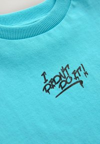 Tirkīza krāsas kokvilnas treniņjakas ar ribotu apaļu kakla izgriezumu un melnu tekstu "ES TO NEIZDARĪJU!" graffiti stila dizainā.