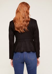 Svart fuskmockajacka med peplumskärning, långa ärmar och en slät textur, tillsammans med blå denimjeans med bakfickor.