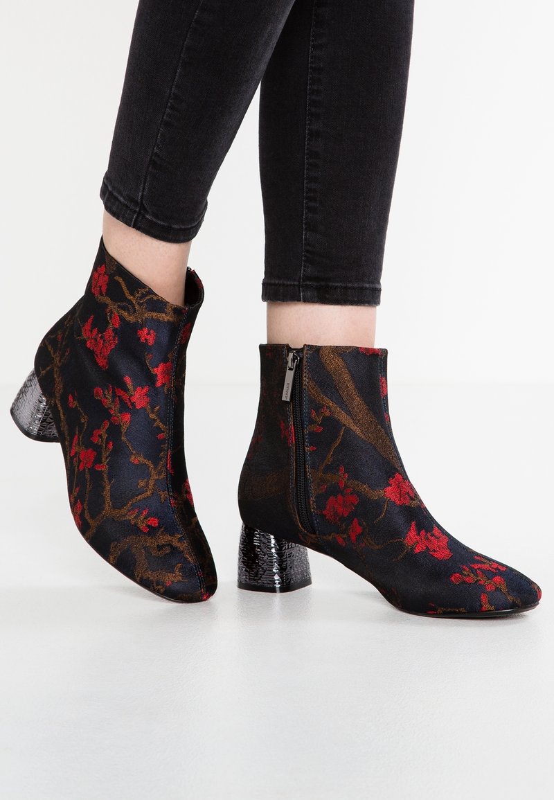 Bottes noires à la cheville avec motif floral rouge, talons épais texturés et fermeture éclair latérale portées avec un pantalon noir ajusté.