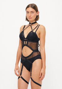 Bodysuit noir avec maille transparente, bonnets structurés et détails de bretelles décoratives. Comprend des sangles de jarretelles ajustables et des accents en découpe.