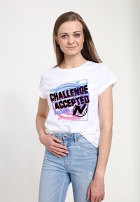 Hvid bomulds t-shirt med et iøjnefaldende, flerfarvet trykt design med teksten "Challenge Accepted" og et logo, parret med lyseblå jeans.