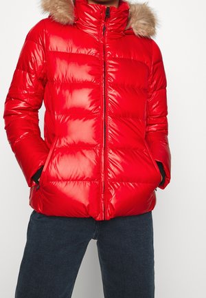 Persona con chaqueta acolchada roja brillante con capucha con borde de piel y manos en los bolsillos, combinada con vaqueros azul oscuro.