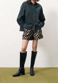 Camicia oversize verde scuro, pantaloni corti beige a fantasia con design floreale nero e stivali neri con tacco al ginocchio, su uno sfondo neutro.