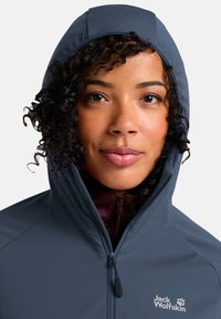 Giacca a cappuccio blu scuro con una texture liscia, zip frontale e logo sul petto. Presenta un design aderente e colletto alto.