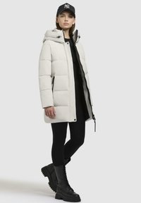 Giacca a vento in beige chiaro con cappuccio, dotata di zip anteriore e dettagli neri. Abbinata a leggings neri e stivali combat con lacci.