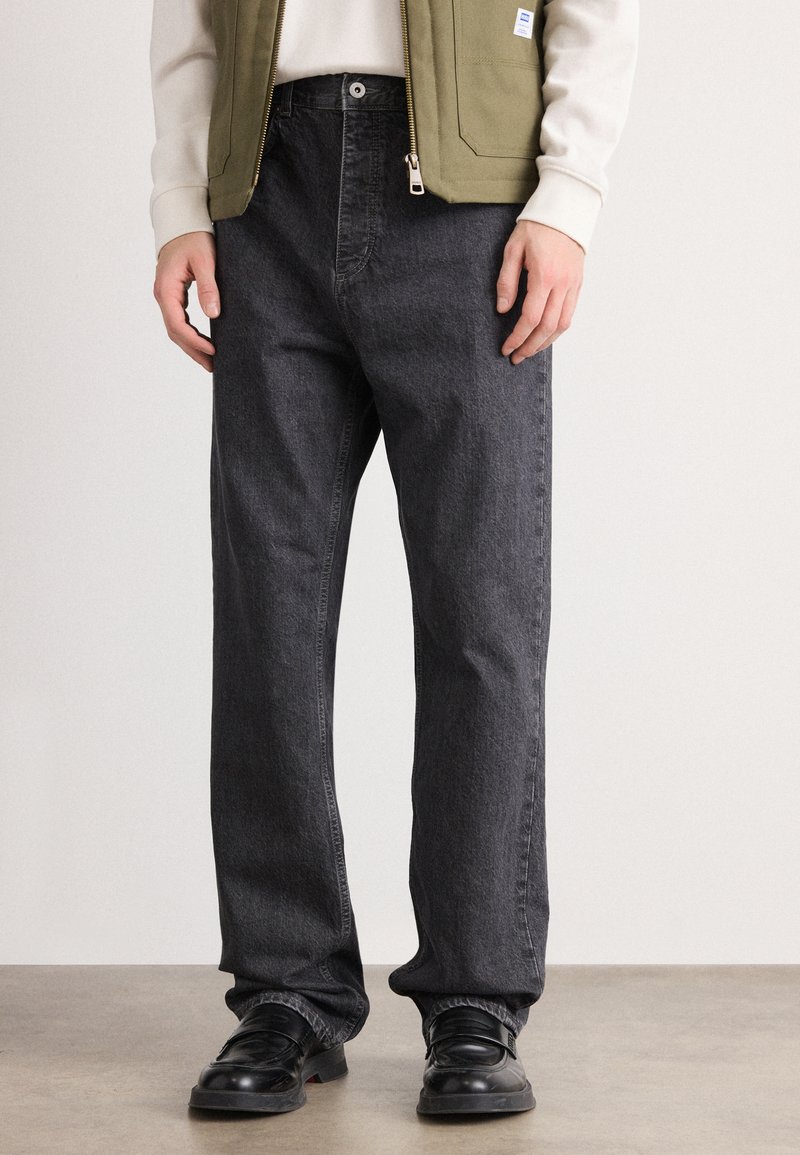 Nederste del af en person iført løse sorte denim jeans, sorte lædersko, en olivengrøn vest og en lysekræm langærmet skjorte.