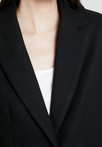 Detailní záběr osoby nosící černé strukturované sako přes bílé žebrované tričko, zobrazující detaily výstřihu a límce.