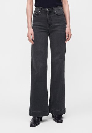 Person iført sort jeans med høj talje og vide ben samt sorte hælede sko, stående mod en ensfarvet lys baggrund.