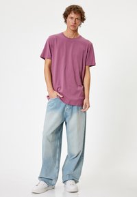 T-shirt viola a maniche corte abbinato a jeans azzurri larghi e sneaker bianchi, in piedi contro uno sfondo neutro.