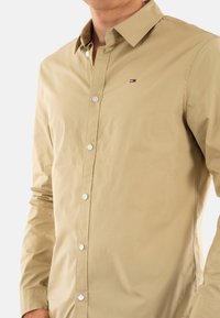 Camicia beige a maniche lunghe in cotone, con colletto button-down, bottoni bianchi e un piccolo logo vicino all'area del petto.