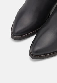 Botas de cuero negro hasta el tobillo con puntas afiladas y detalles de costura visibles. Textura suave y diseño minimalista.