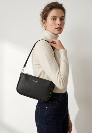 Donna con dolcevita color crema e jeans scuri che porta una piccola borsa a tracolla nera con cerniera e tracolla argentate.