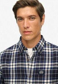 Niebieska i biała koszula w kratę z kołnierzykiem zapinanym na guziki, z brązowymi guzikami i naszywką z logo na kieszeni piersiowej. Materiał bawełniany.