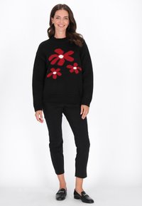 Schwarzer Strickpullover mit rotem Blumenmuster, der drei Blumen mit weißen Zentren zeigt. Kombiniert mit schwarzen schmalen Hosen und Slippern.