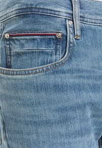 Los jeans de mezclilla de color azul claro presentan un bolsillo trasero con acentos de costura en rojo y azul. La tela tiene un acabado suave y texturizado con zonas desgastadas.