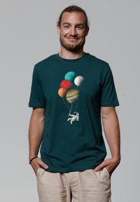 T-shirt en coton bleu-vert foncé arborant un graphique fantaisiste d'un astronaute tenant des ballons en forme de planète de différentes teintes.