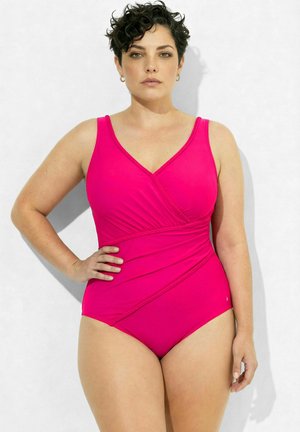 Kobieta z krótkimi kręconymi włosami, nosząca jaskraworóżowy jednoczęściowy strój kąpielowy, stoi z jedną ręką na biodrze na jednolitym jasnym tle.