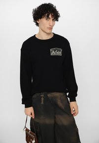 Sort strikket sweater med ribbet tekstur og et beige broderet "Aries" logo på brystet, parret med brune cargo bukser og en lille håndtaske.