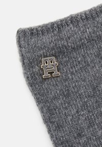 Grijze gebreide beanie met een omgeslagen rand en een kleine, metalen logo pin in goud en donkergrijs. Zachte textuur en warm ontwerp.