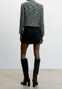 Veste en tweed tissé vert, mini-jupe noire et bottes noires montantes jusqu'aux genoux. Texture lisse, coupe ajustée, manches longues, détails minimalistes.