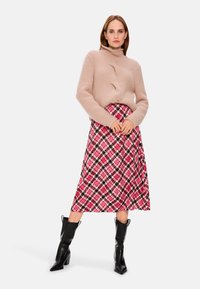 Beige Strickpullover mit hohem Kragen, kombiniert mit einem rosa gemusterten Midirock mit Schwarz- und Weißakzenten. Schwarze Absatzstiefel.