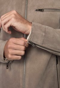 Chaqueta de ante color tostado con cremallera en la parte delantera. Cuenta con un bolsillo con cremallera en el pecho y puños con cremallera. Textura suave y diseño aerodinámico.
