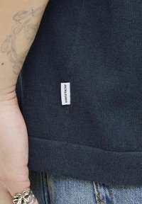 Námorně modrý pletený svetr s texturedním povrchem, na dolním lemu s malou bílou etiketou s černým textem. Jemný detail na manžetách.
