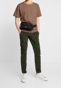 Czarna nerka z logo, noszona na brązowo-beżowej koszulce w paski. Dopełnieniem stroju są oliwkowe spodnie i białe trampki.