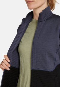 Chaqueta de cremallera en azul marino y negro con cuello alto y paneles texturizados, llevada sobre una camisa verde claro; detalles de costura visibles.