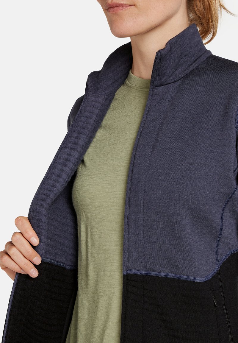 Marineblaues und schwarzes Zip-Jackett mit hohem Kragen und strukturiertem Besatz, getragen über einem hellgrünen Hemd; sichtbare Steppdetails.