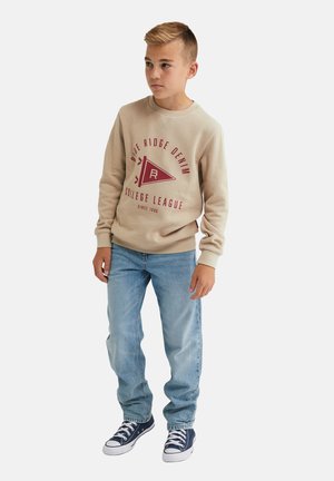 Beige sweatshirt met rode grafische ontwerpen en tekst, gecombineerd met lichtblauwe jeans en marineblauwe sneakers. De sweatshirt heeft ribgebreide manchetten en een zoom.