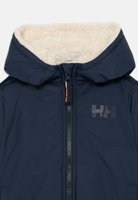 Giacca blu navy con cappuccio foderato in pelliccia, zip frontale e dettaglio del logo nero. Materiale esterno liscio, texture imbottita e design pratico.